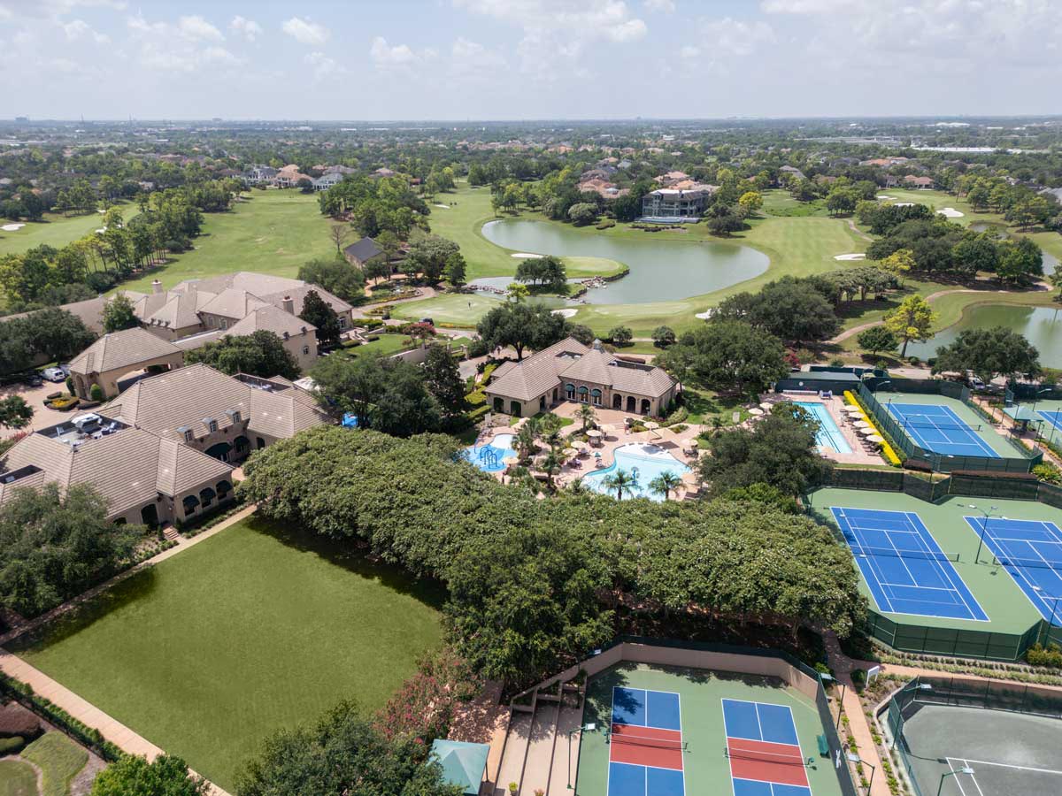 Aerial view of a residential area featuring tennis courts, a swimming pool, clubhouse, green lawns, and a golf course with lakes.