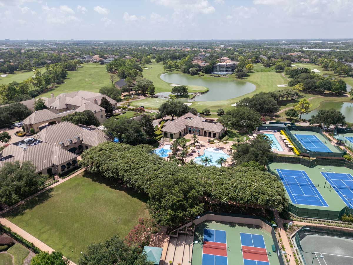 Aerial view of a residential area featuring tennis courts, a swimming pool, clubhouse, green lawns, and a golf course with lakes.