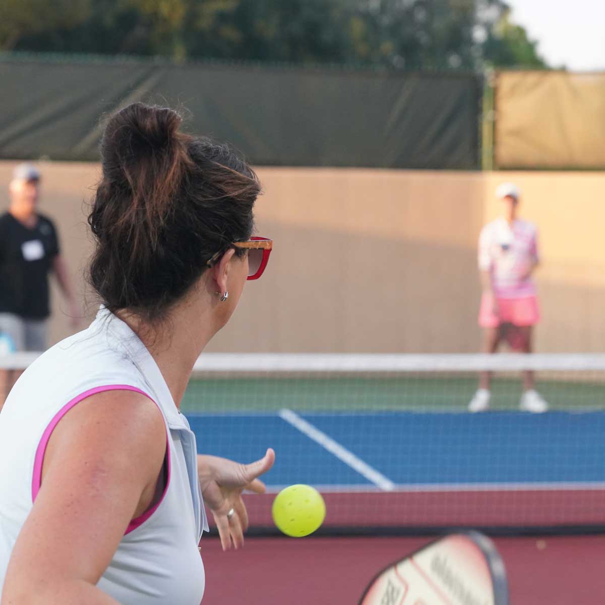 Woman preparing to hit a yellow pickleball on an outdoor court with two players in the background.