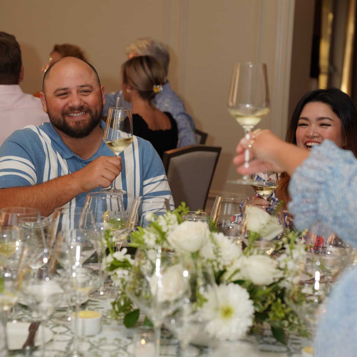 Smiling man and woman clinking white wine glasses at a decorated dining table with white flowers.