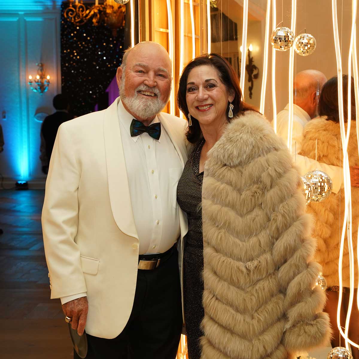 An older man in a white tuxedo jacket and bow tie standing next to a woman wearing a fur coat and black dress, both smiling at a formal event.