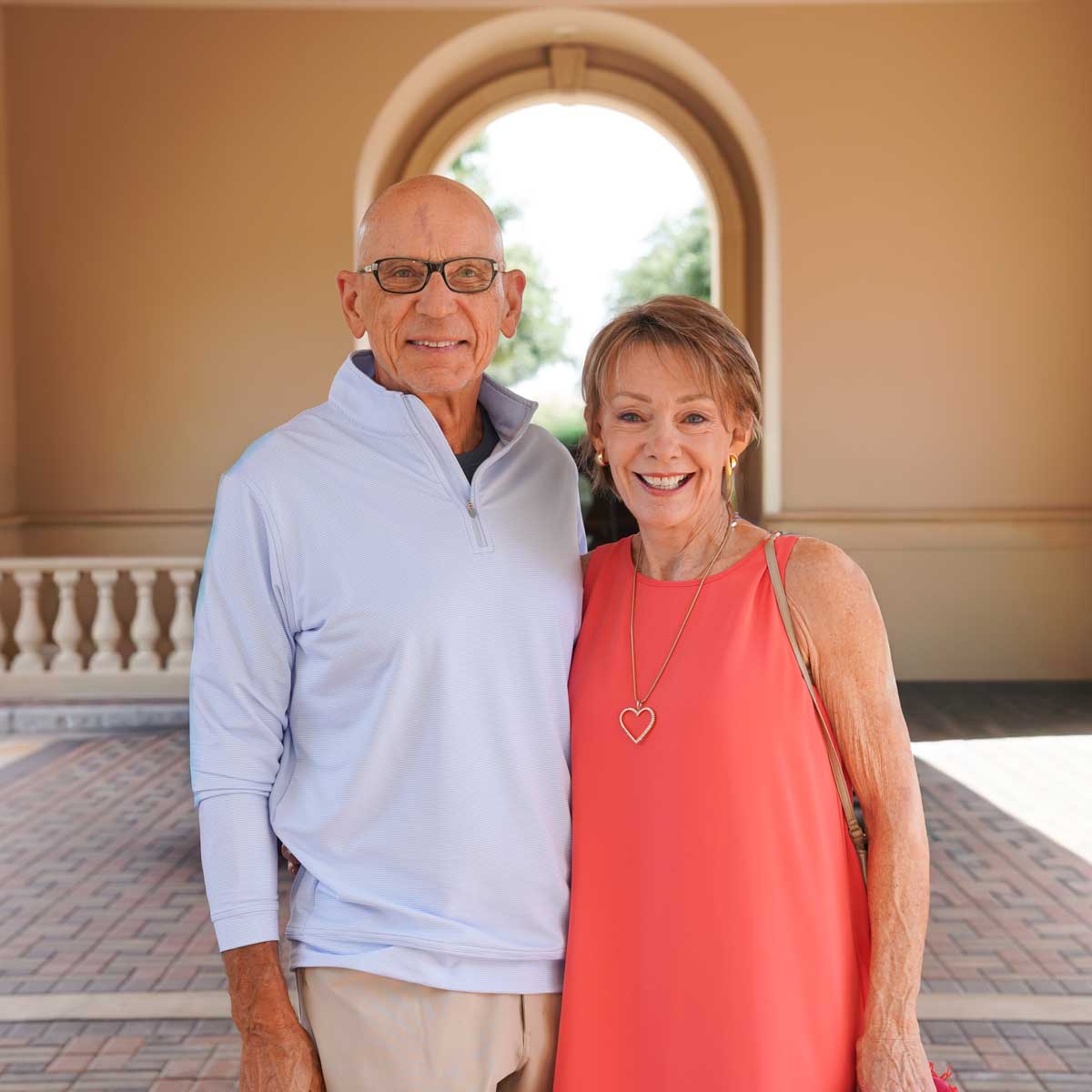 Smiling elderly couple standing close together under an archway with a tiled floor.