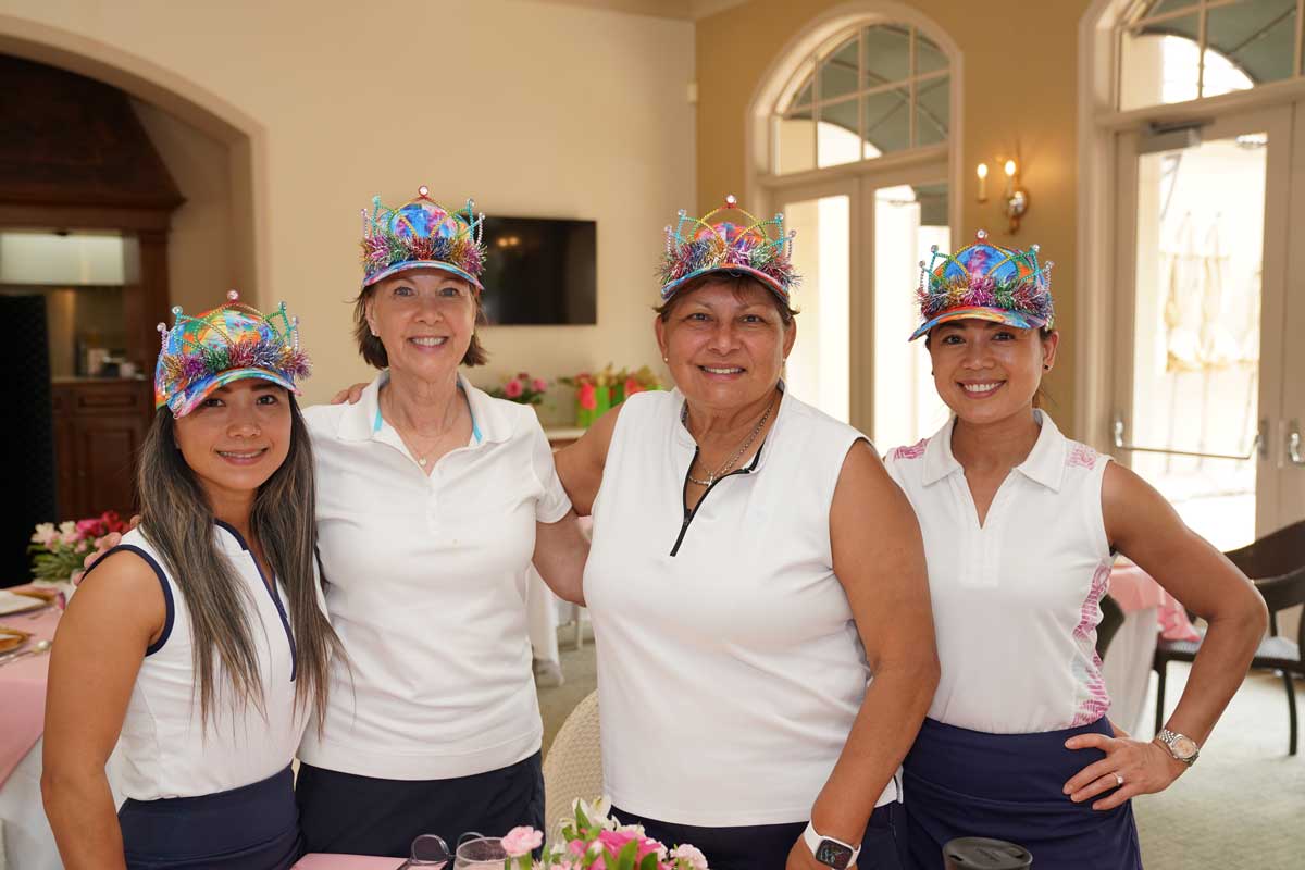 Four smiling women indoors wearing colorful decorated caps and white golf shirts.