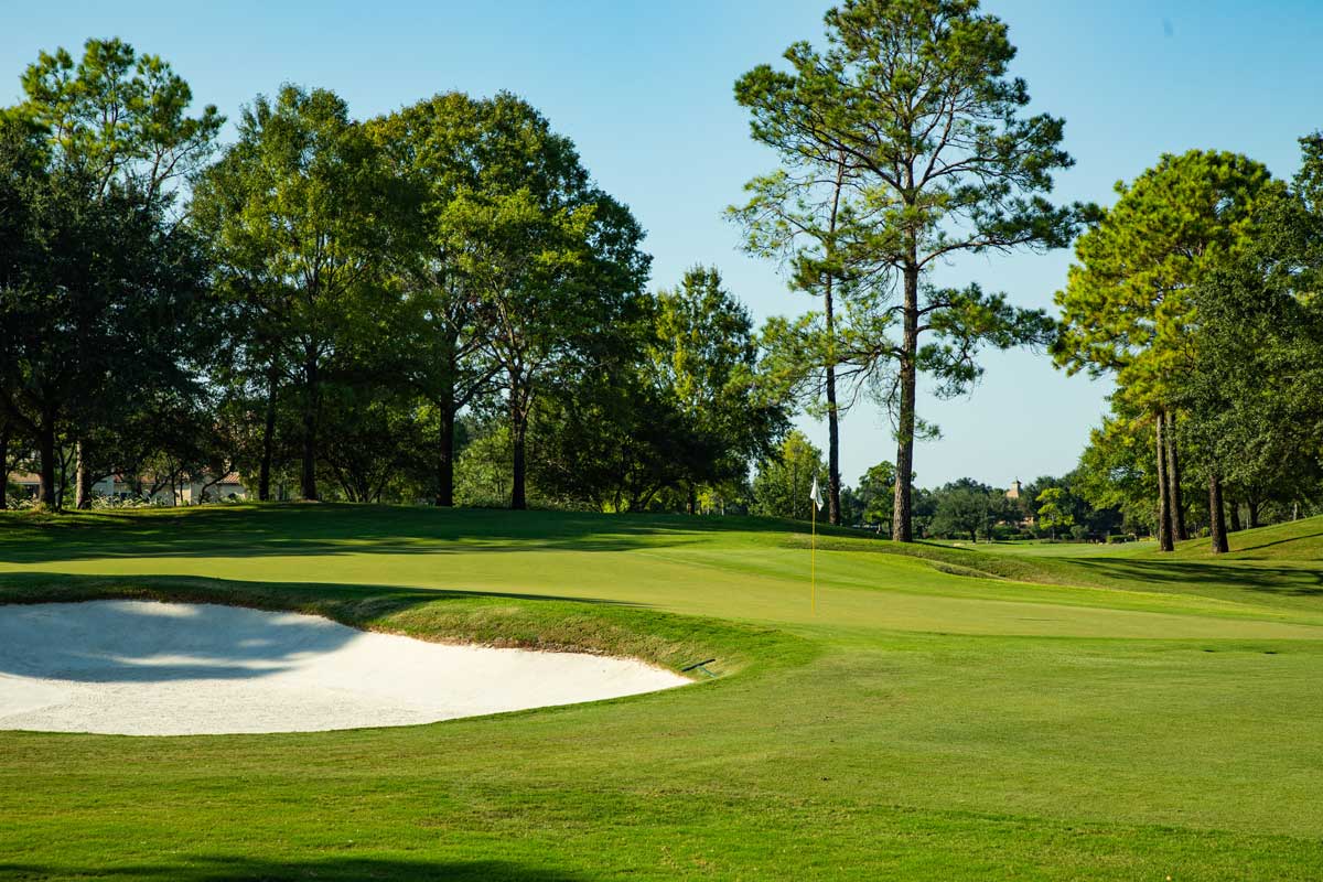 Golf course green with a sand bunker on the left and trees bordering the fairway under a clear blue sky.