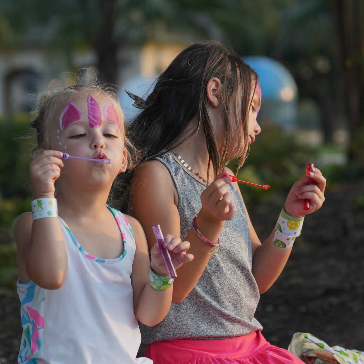 Two young girls with face paint blowing bubbles outdoors in the evening light.