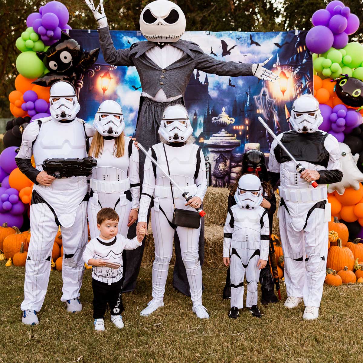 Group of people dressed in Star Wars stormtrooper costumes and a character with a large skeleton head in a spooky Halloween-themed setting with pumpkins and balloon decorations.