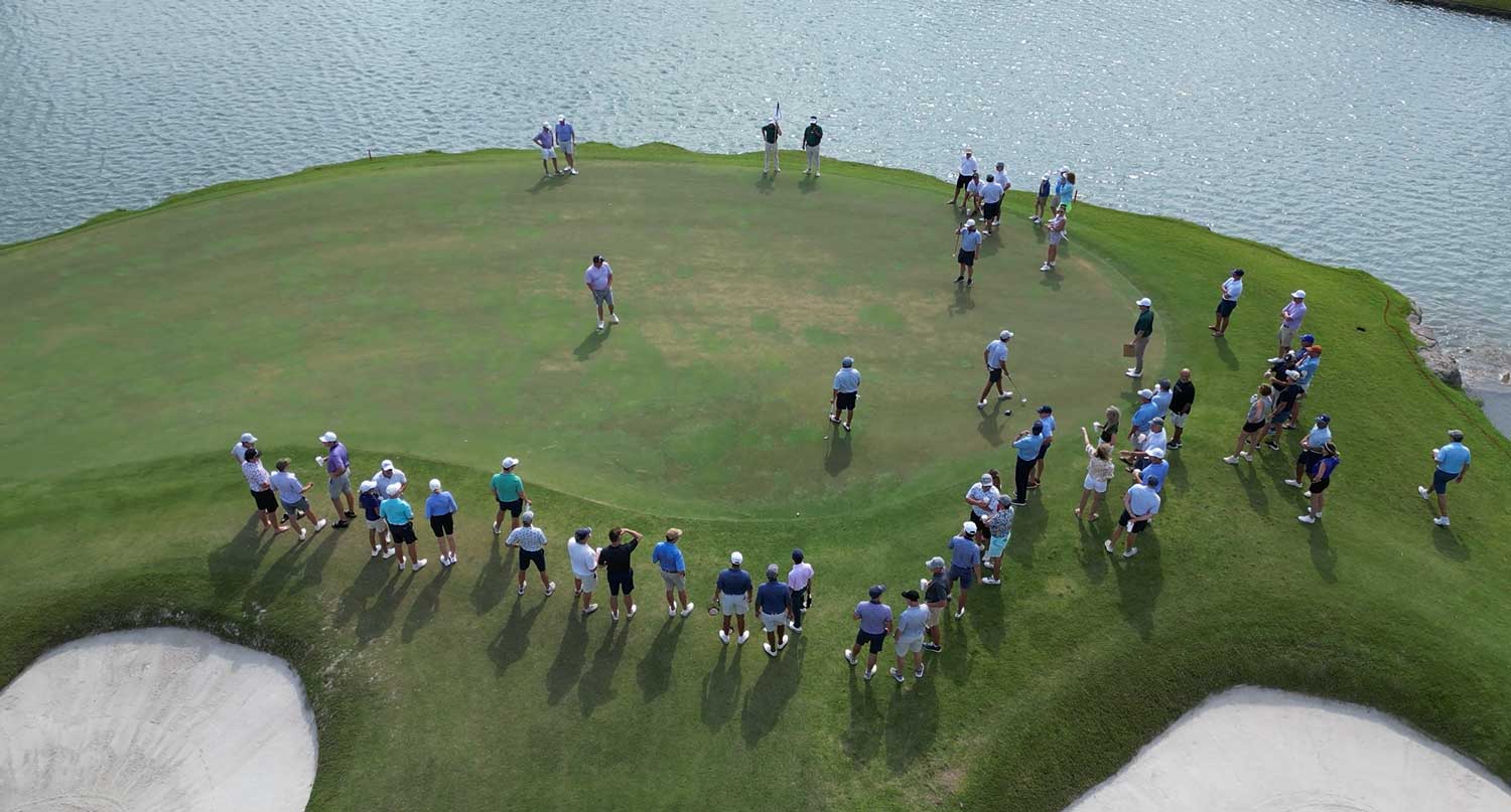 Aerial view of a golf green surrounded by spectators watching players putting near a water hazard and sand bunkers.