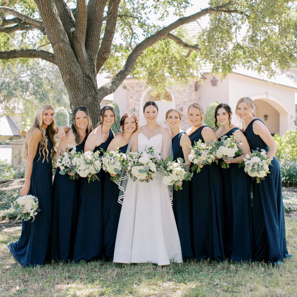 Bride in white gown and veil standing outdoors with eight bridesmaids in navy dresses holding white and green floral bouquets under a large tree.