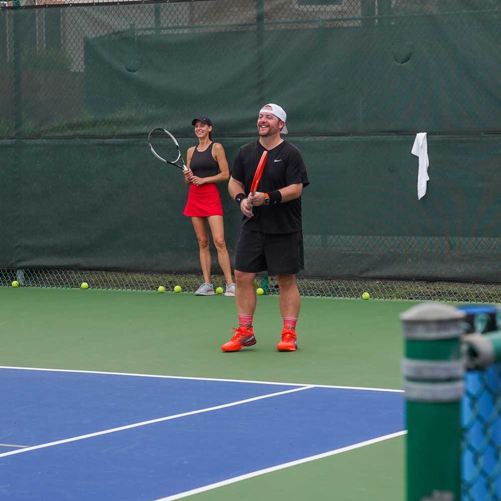 Man and woman on tennis court holding racquets and smiling, with tennis balls scattered along the fence.