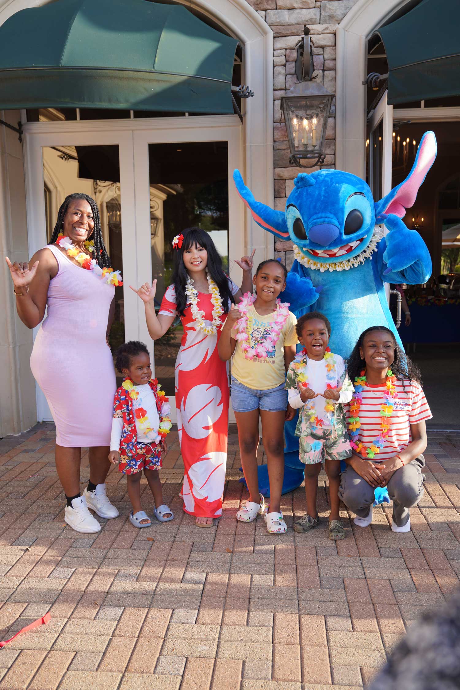 Group of children and adults wearing colorful leis posing with a person in a large blue Stitch costume outdoors in front of a building.