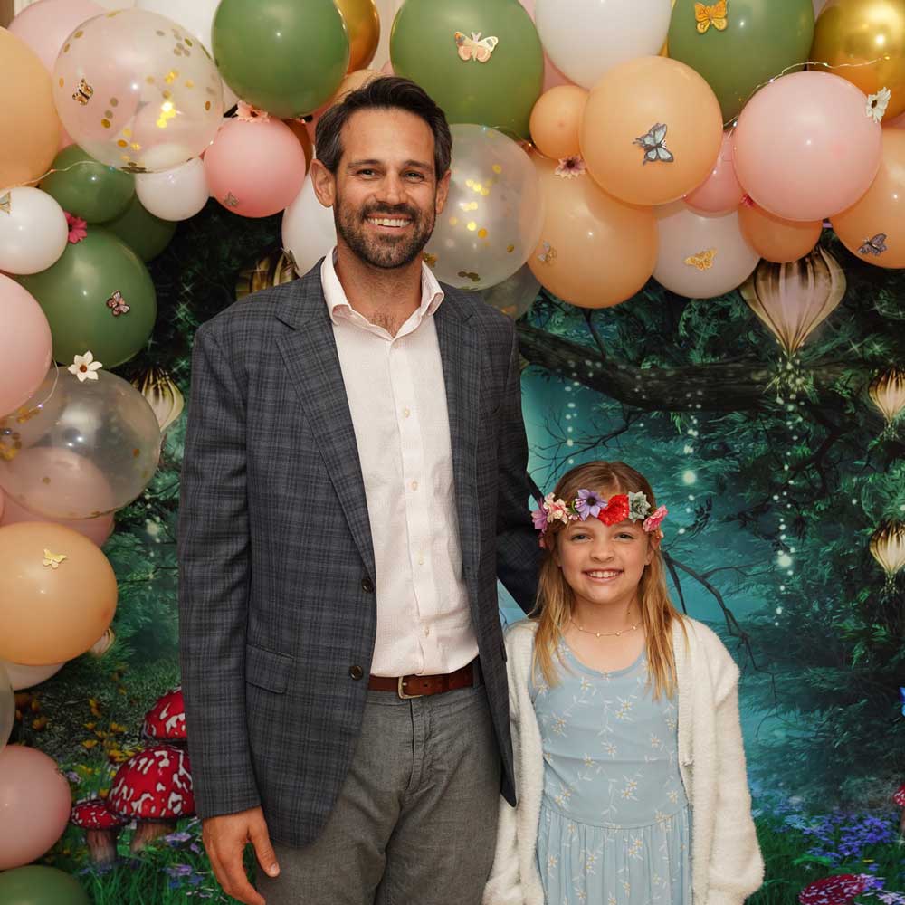Smiling man in a gray checkered blazer and young girl wearing a blue dress and flower crown standing in front of a balloon arch and fantasy forest backdrop.