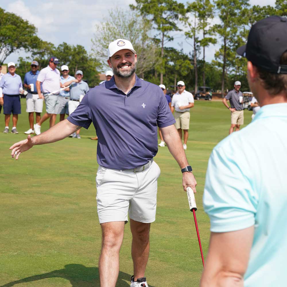 Smiling man in a purple shirt and white cap extending his arms on a golf course with other golfers in the background.