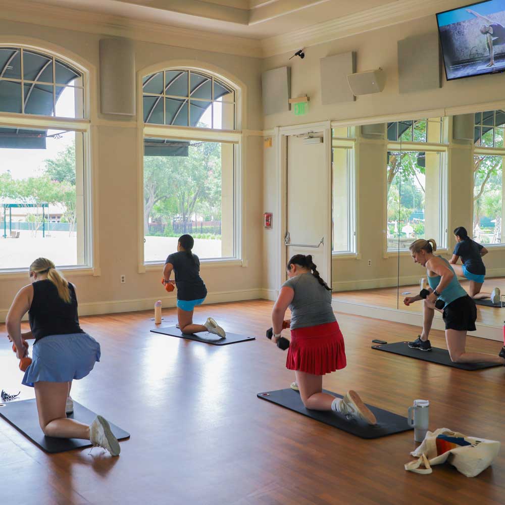 Four women exercising with dumbbells on mats in a bright fitness studio with large windows and mirrors.
