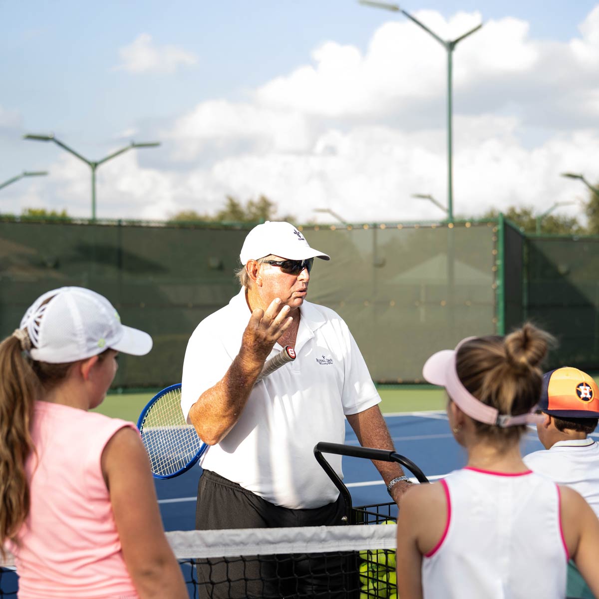 Tennis coach in white shirt and cap giving instructions to children on a tennis court.
