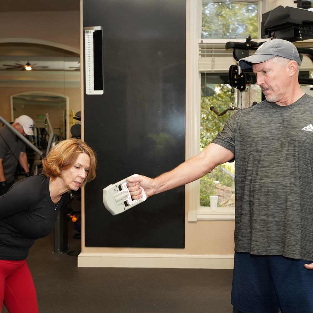 A man in a gray shirt and cap holds a digital hand dynamometer while a woman in black and red watches attentively in a gym setting.