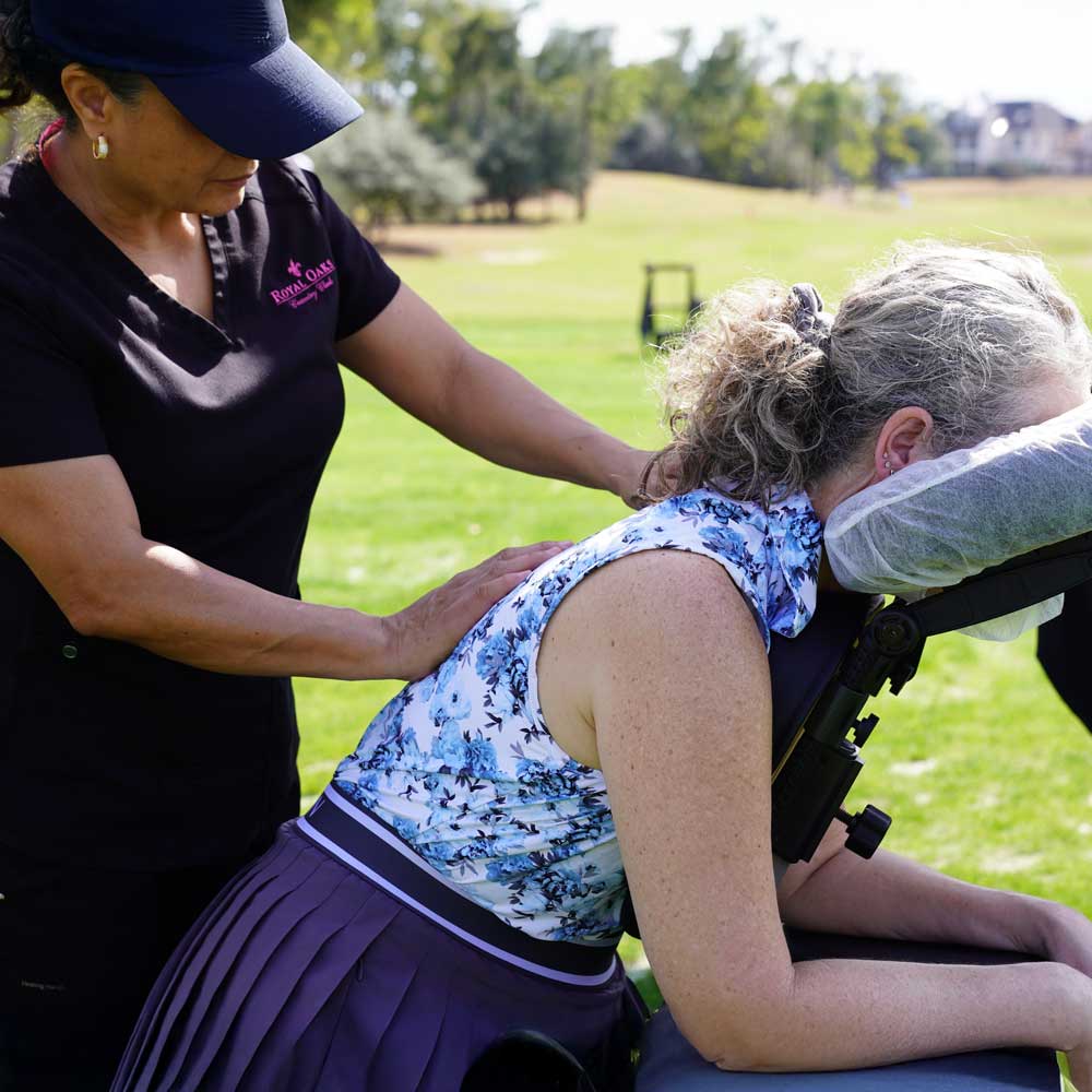 A woman receiving an outdoor seated massage on her upper back while resting her head on a cushioned massage chair.