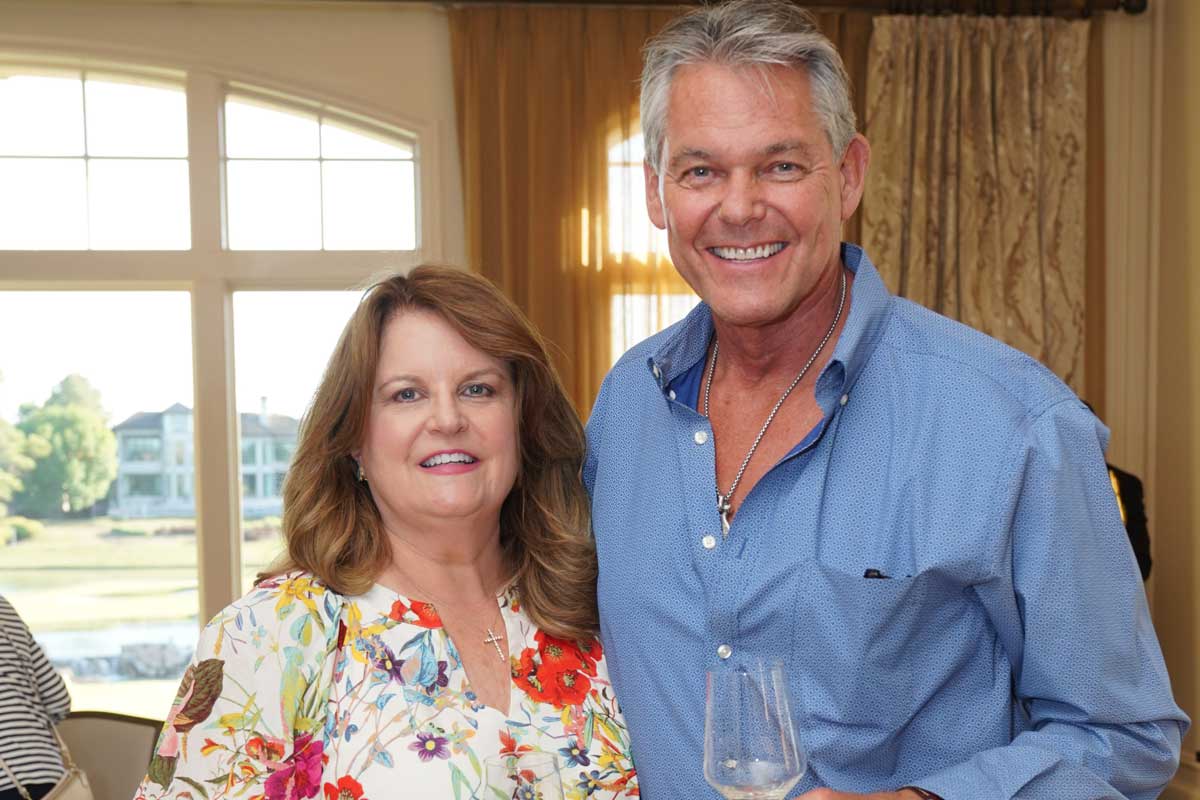Smiling middle-aged couple indoors, the woman wearing a floral blouse and the man a blue shirt holding a wine glass.