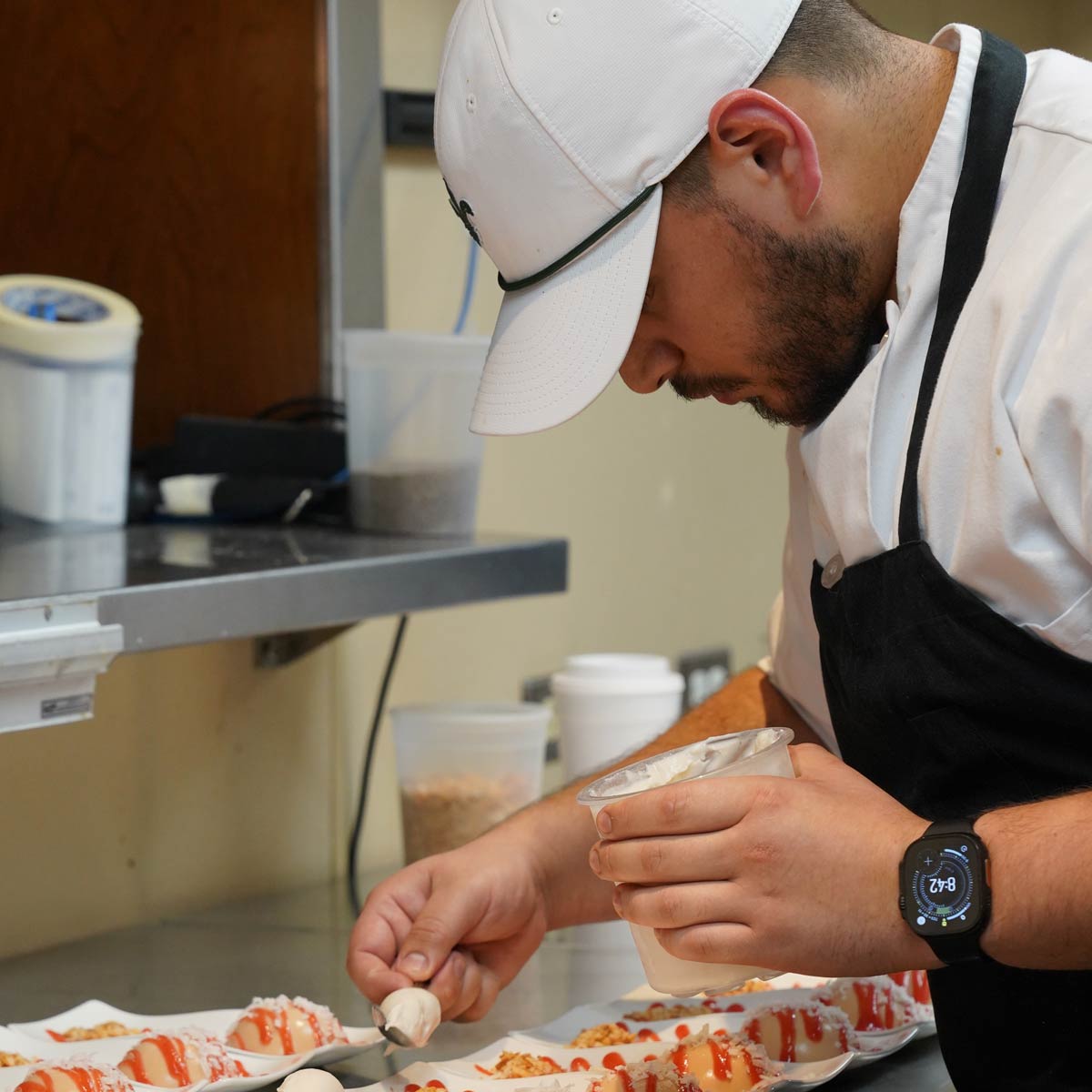 Chef wearing a white cap and black apron meticulously garnishing plated desserts with a cream from a small container.