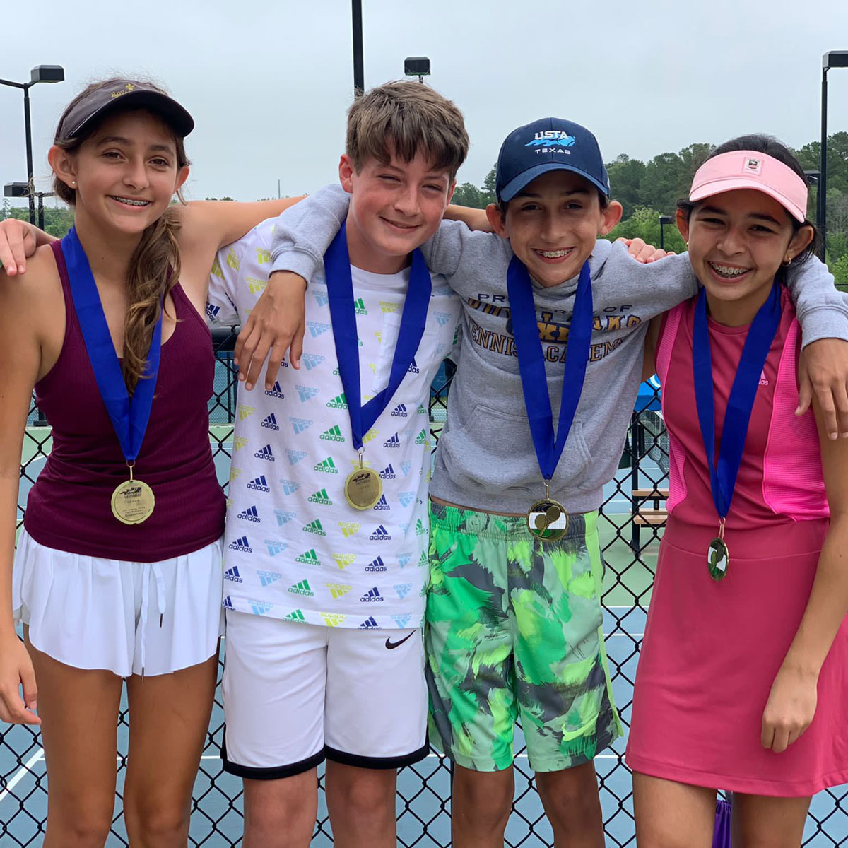 Four smiling children wearing medals around their necks stand arm in arm on a tennis court.