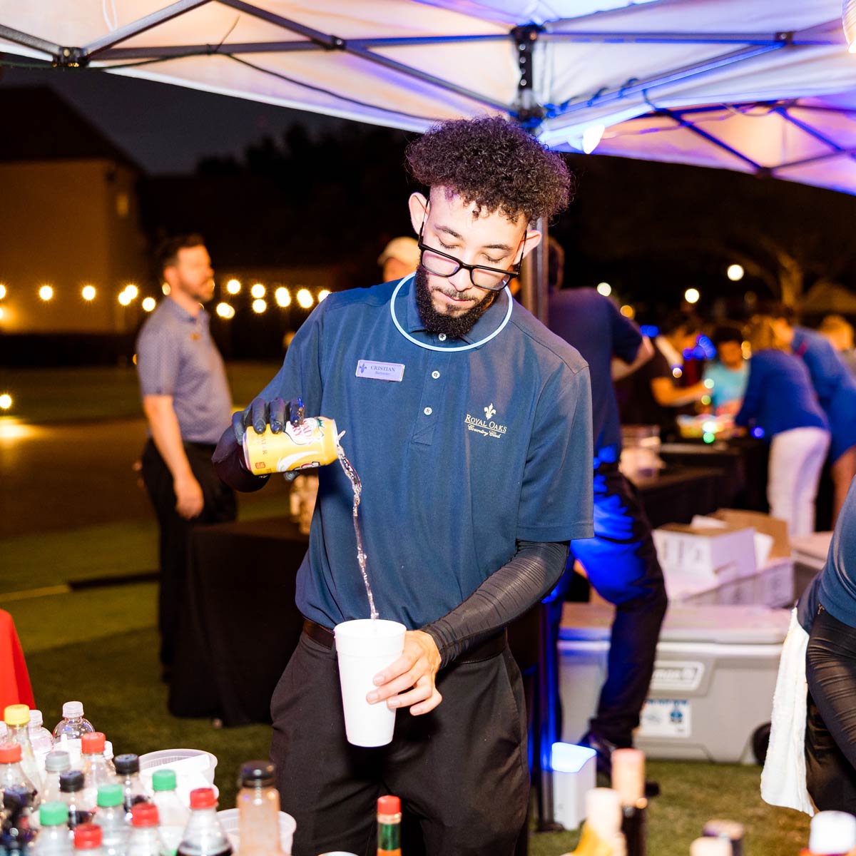 Man wearing glasses and a Royal Oaks Golf Club shirt pouring a drink from a can into a white cup under an outdoor tent at night.