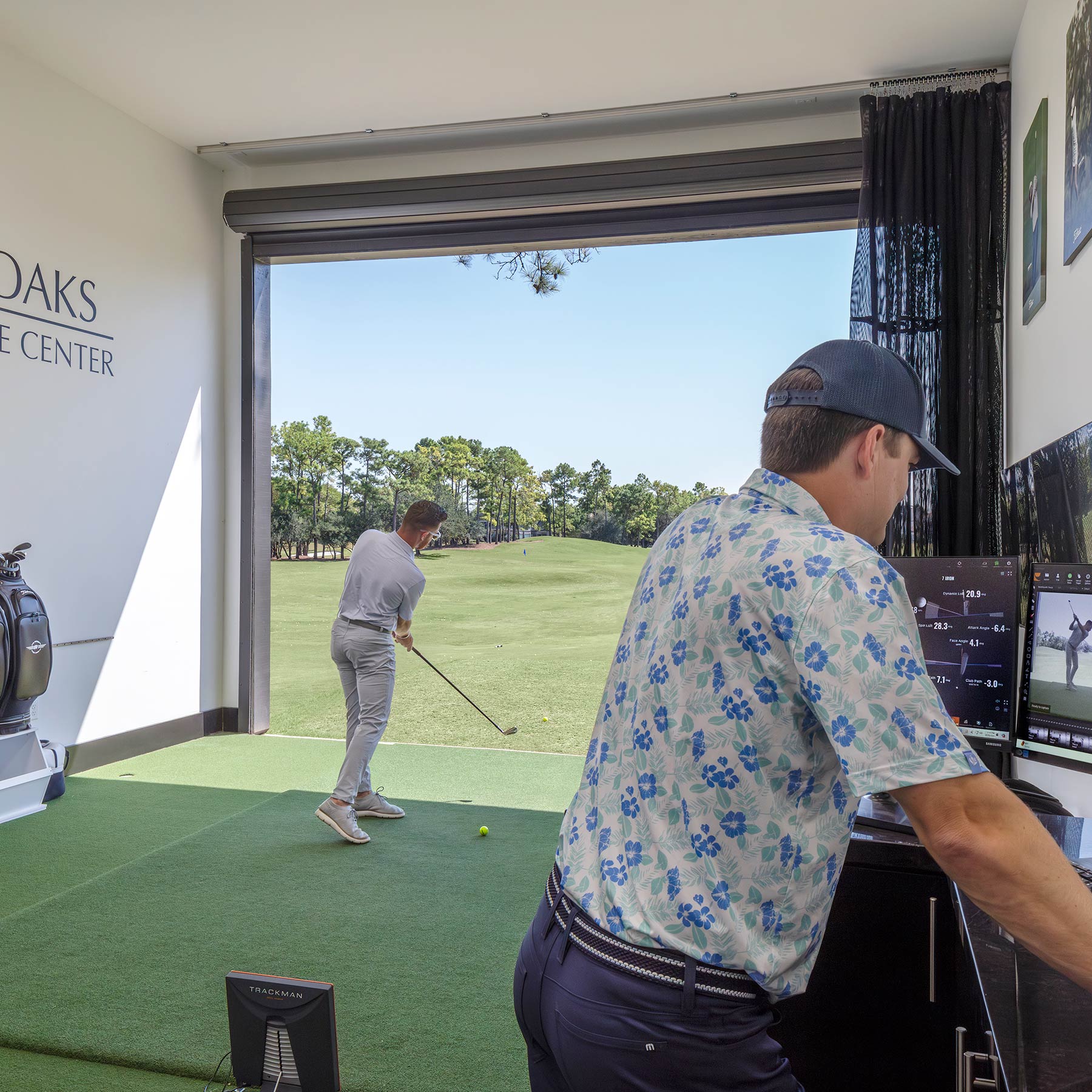 Golf instructor analyzing swing data on screens while student practices golf swing indoors overlooking a green fairway.