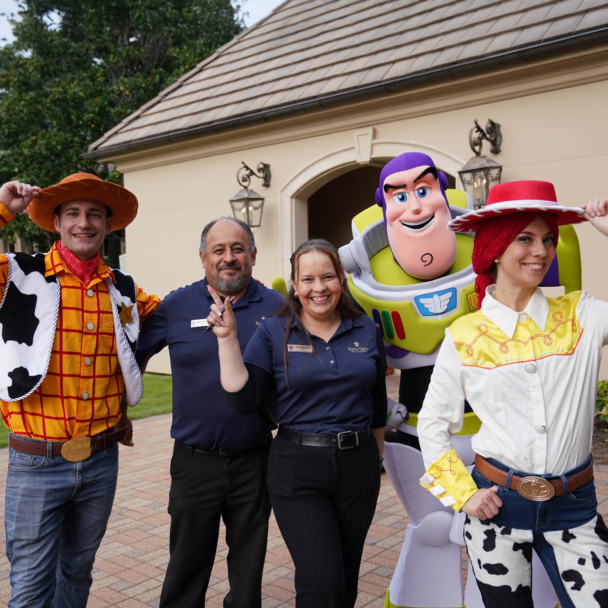 Group of five smiling people, including two dressed as Woody and Jessie and one as Buzz Lightyear from Toy Story, posing outside a building.