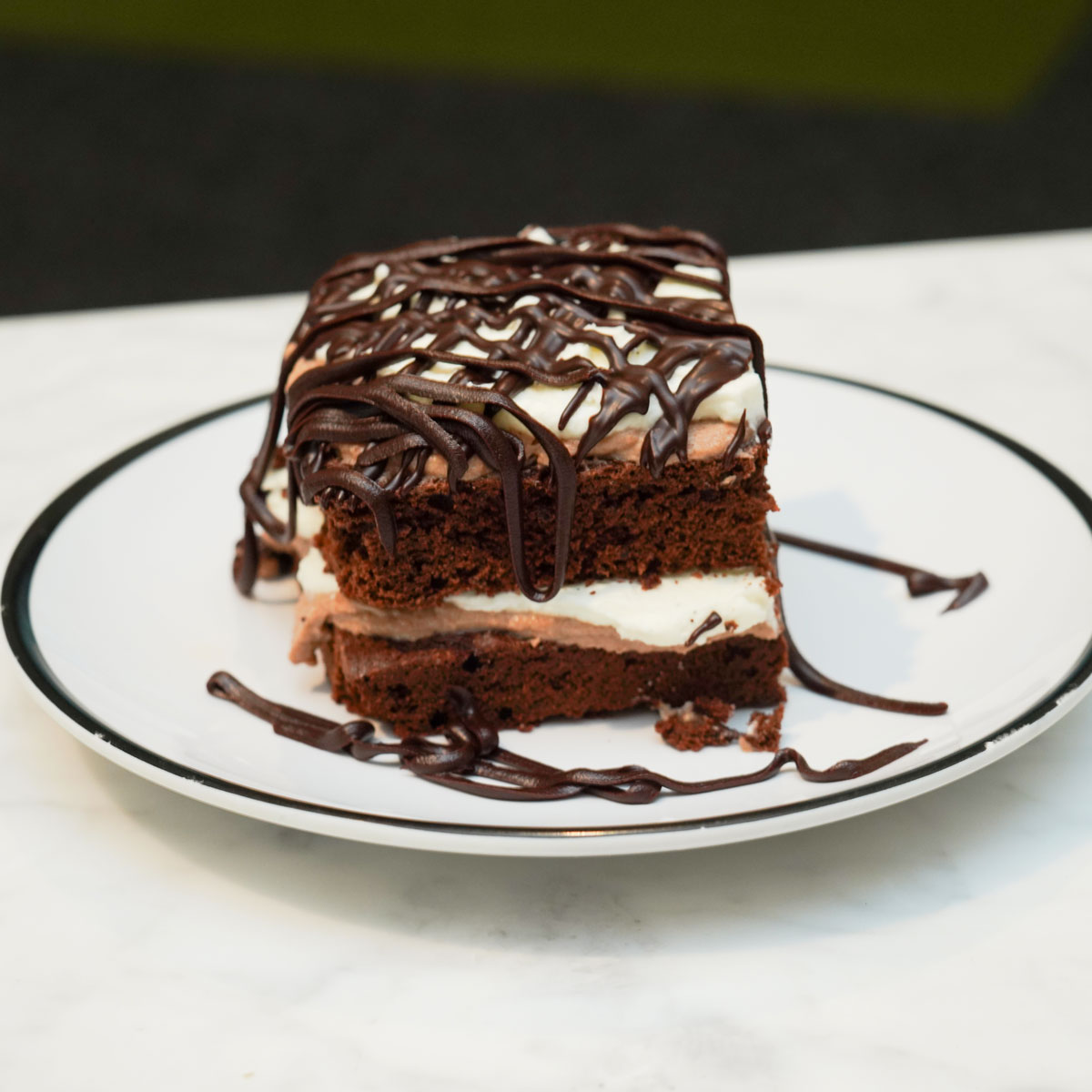 Slice of chocolate cake with white cream layers and chocolate drizzle on a white plate with black rim.