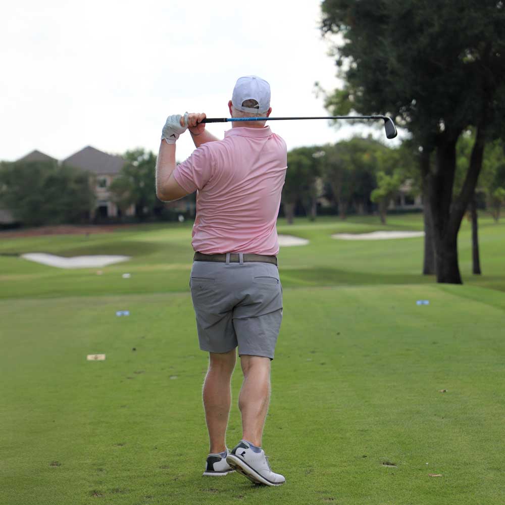 Man in pink shirt and gray shorts swinging a golf club on a golf course.