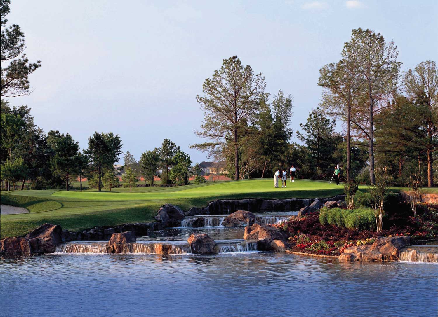 Golfers putting on a green near a cascading rock waterfall by a lake surrounded by trees and landscaping.