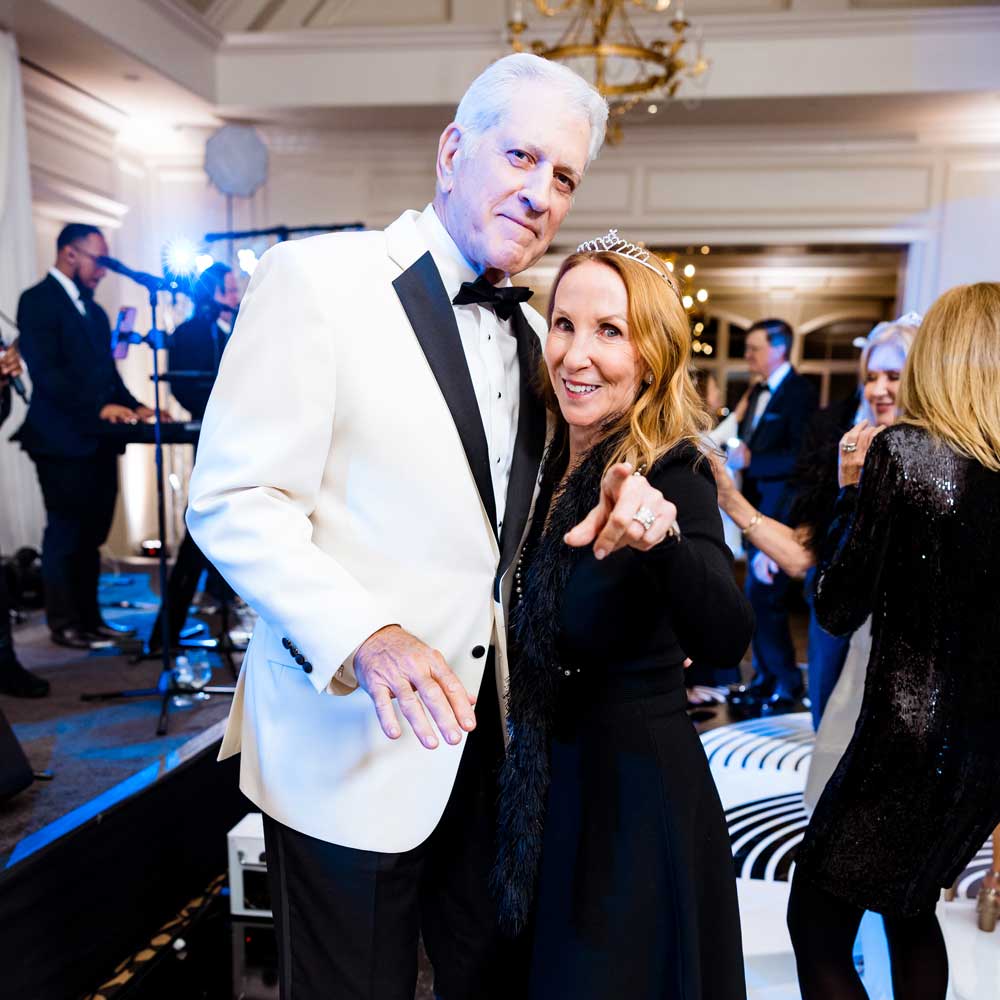 Man in white tuxedo and woman in black dress wearing a tiara, smiling and dancing at a formal indoor event with a live band in the background.