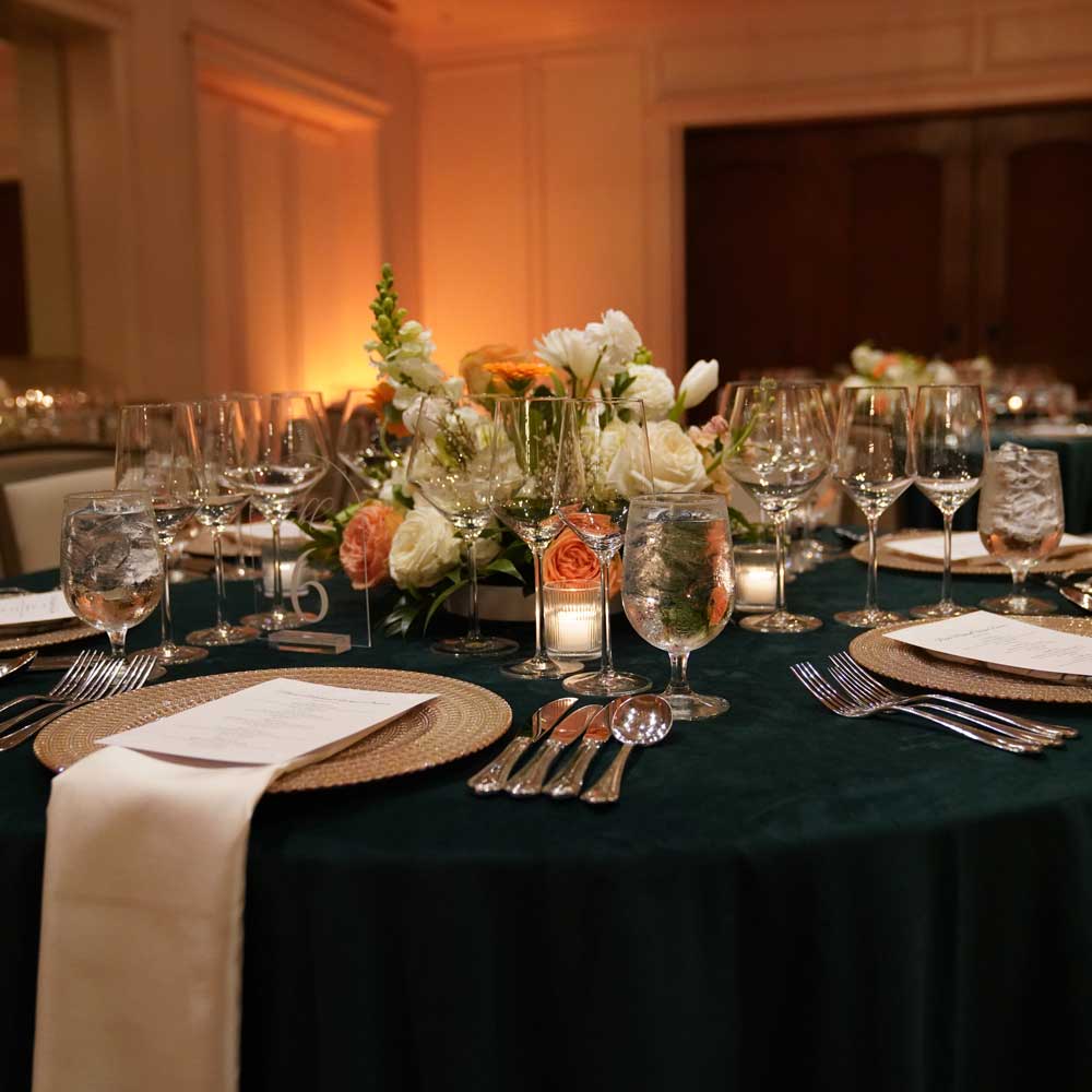 Elegant round banquet table with floral centerpiece, multiple wine glasses, water glasses, cutlery, and menus on woven chargers.