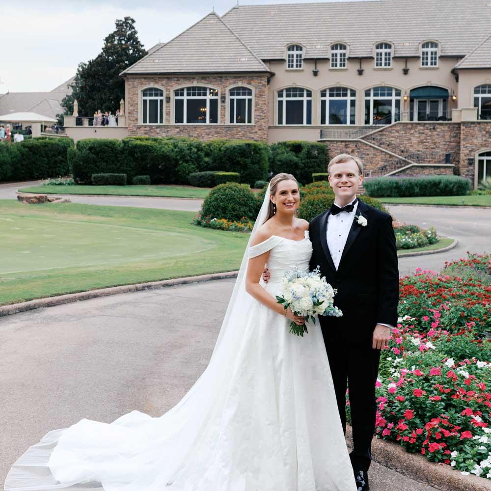 Smiling bride in white gown with veil and bouquet standing next to groom in black tuxedo outside a large stone and brick building with flowers and greenery.