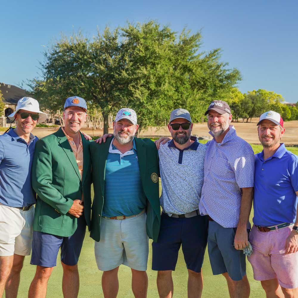 Group of six men standing close together on a golf course, wearing casual golf attire including hats and shorts, with trees and blue sky in the background.