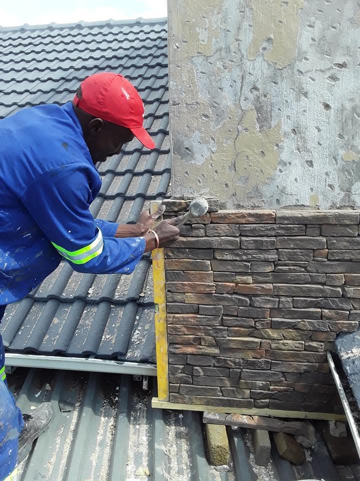 Construction worker installing decorative stone cladding on a wall beside a tiled roof.