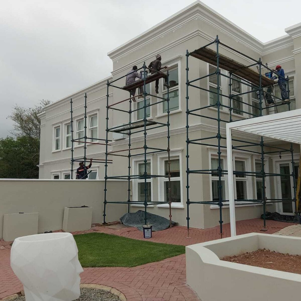 Workers on scaffolding painting or repairing a two-story beige house exterior.