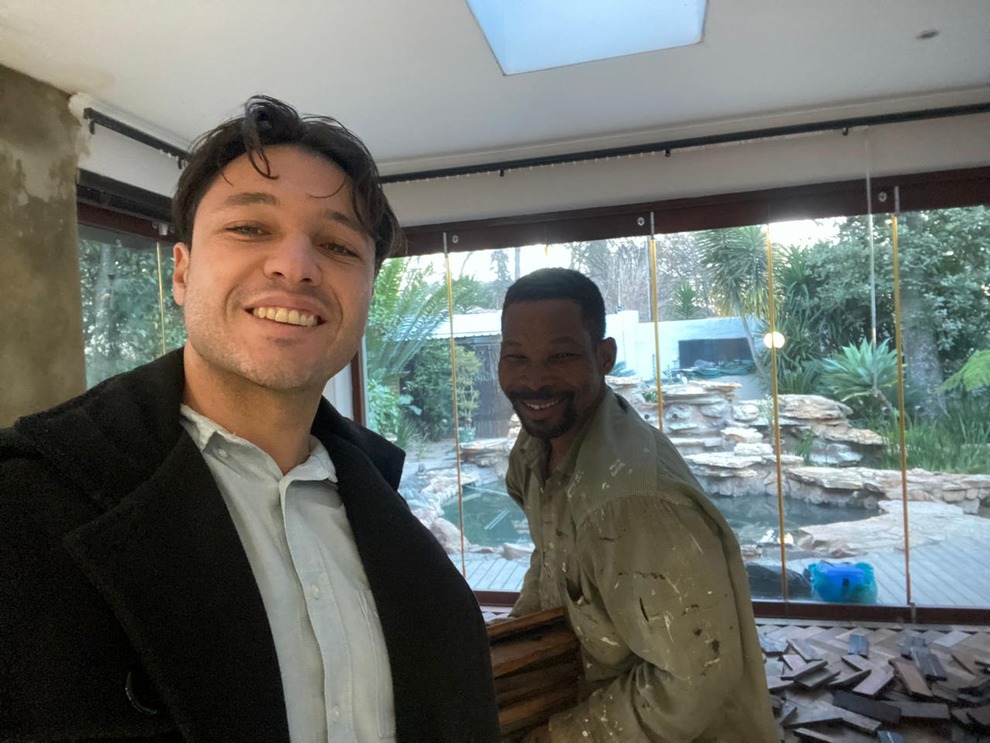 Two men smiling indoors with a large window showing a garden and water feature behind them.