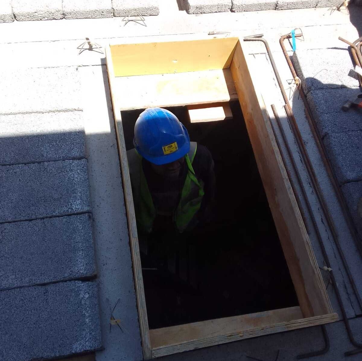 Construction worker with blue hard hat and safety vest in a rectangular floor opening framed with wood.