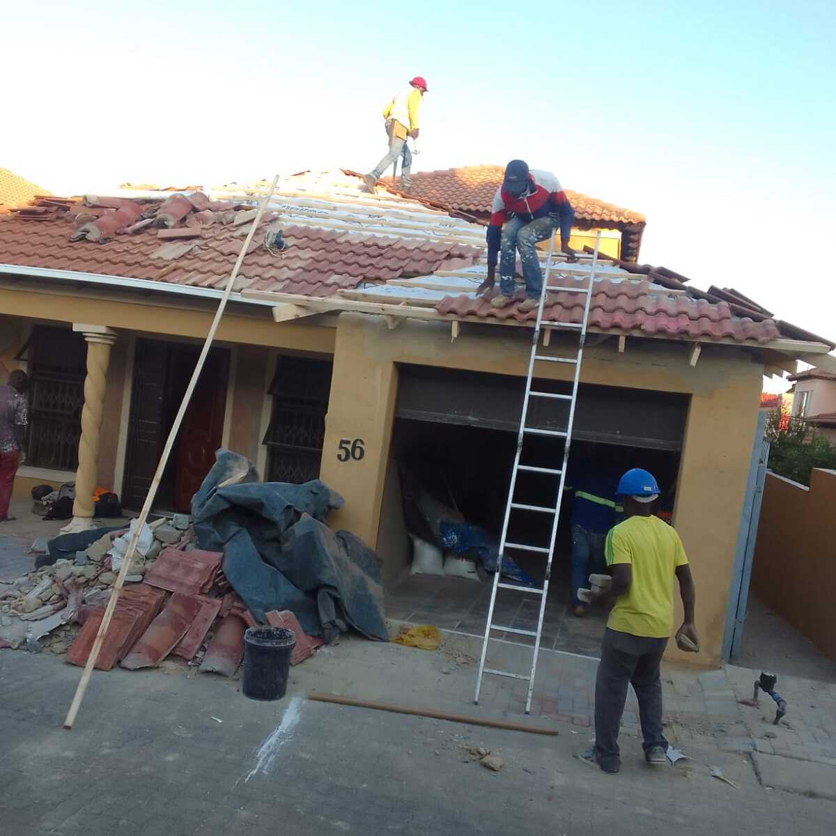 Workers repairing a tiled roof of a yellow house with ladder and construction debris outside.