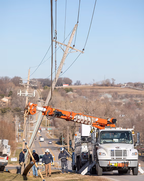 Utility workers repairing a tilted power pole beside a utility truck on a rural roadside.