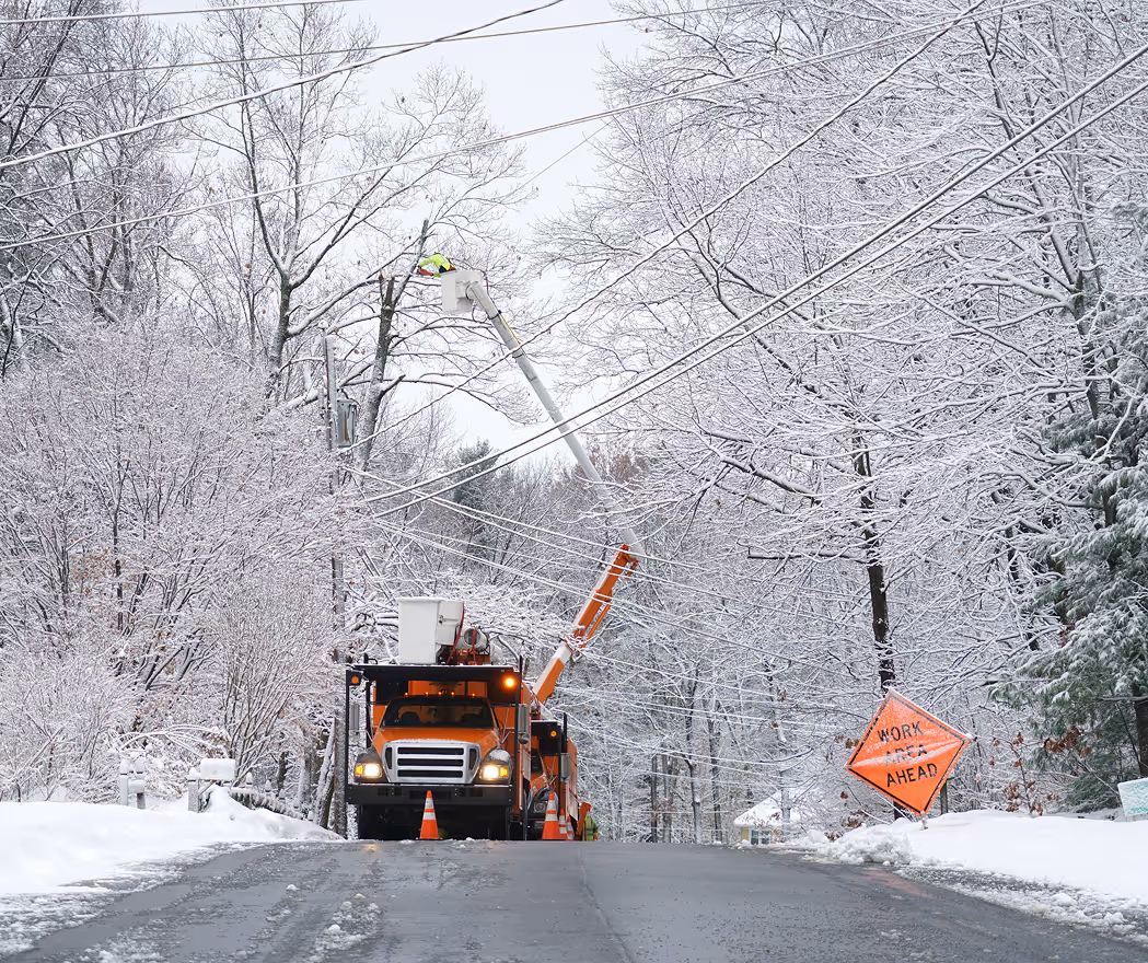 Utility workers in a bucket truck repairing power lines along a snowy road with leafless snow-covered trees and a 'Work Ahead' sign.