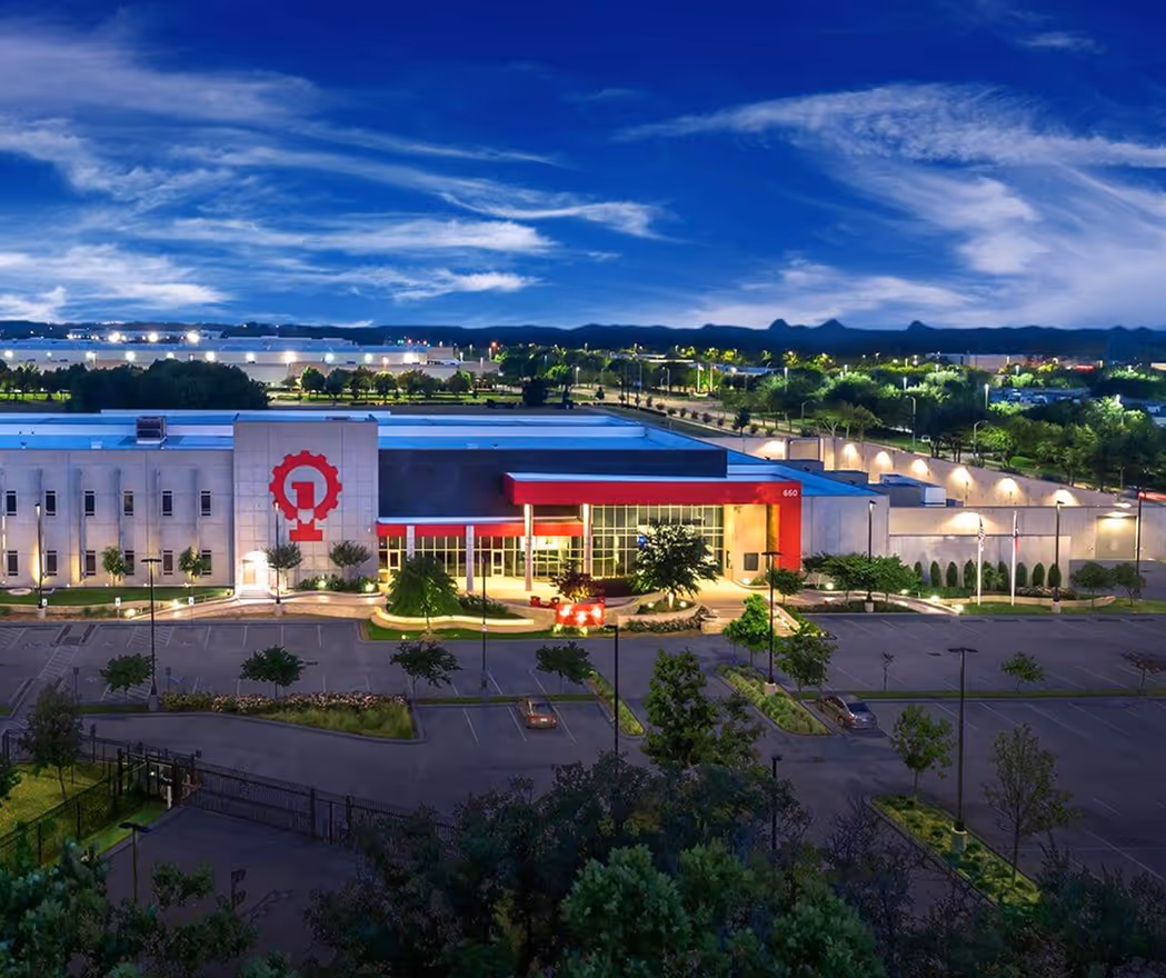 Night view of a modern industrial building with well-lit entrance and red accents, surrounded by trees and parking lots.
