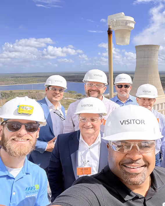 Group of men wearing white visitor hard hats and safety glasses standing outdoors with industrial cooling towers and a water body in the background.