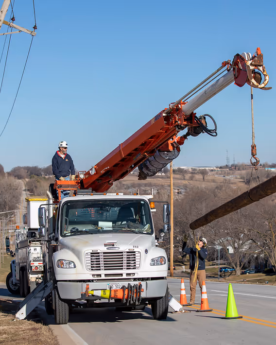 Utility workers operating a crane truck to lift a large wooden utility pole on a street with traffic cones around.