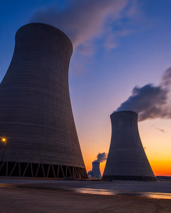 Three large cooling towers emitting steam at a power plant during sunset.