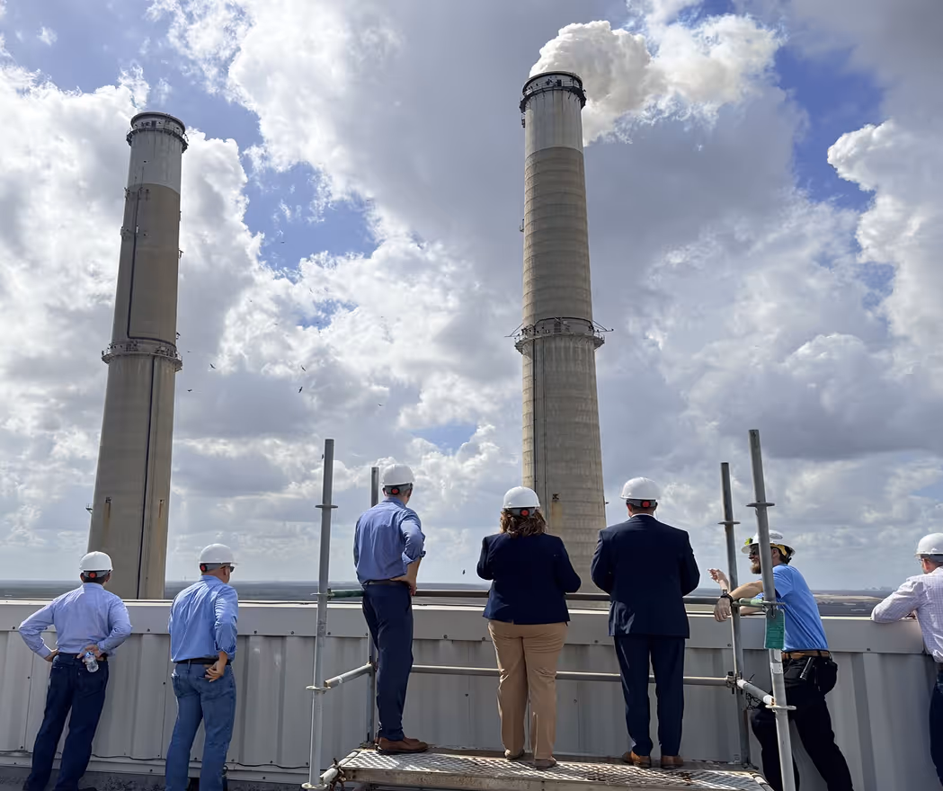 Seven people wearing hard hats and business casual clothing stand on a rooftop platform overlooking two tall smokestacks emitting white smoke under a partly cloudy sky.