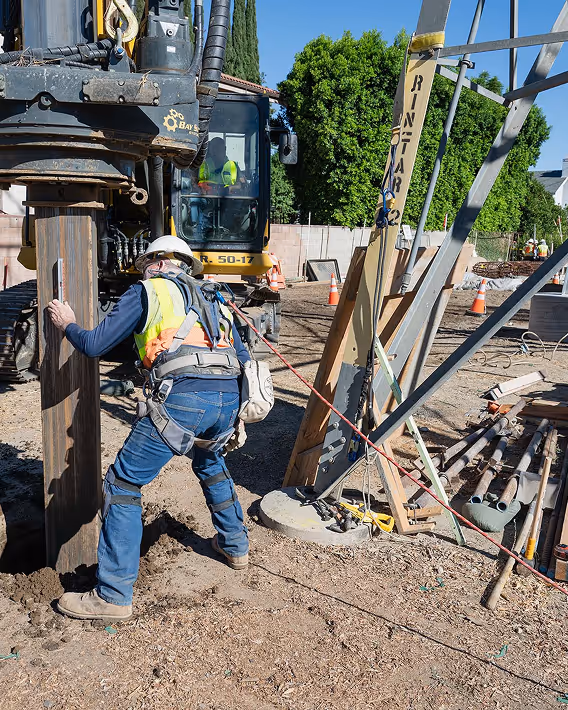 Construction worker in safety gear guiding a pile driver machine during a foundation installation on a sunny site.