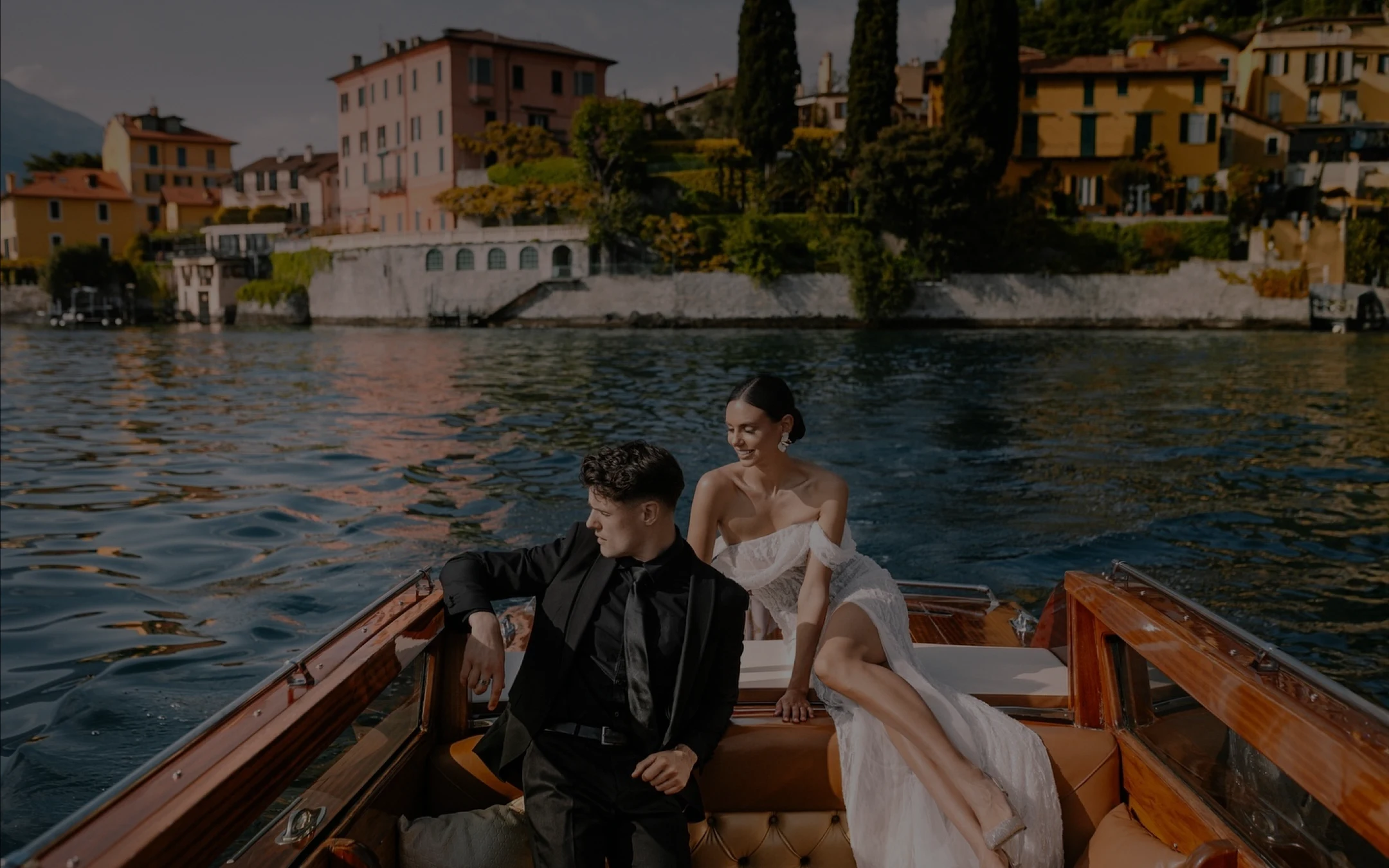 Elegant couple dressed in black suit and white dress sitting on a wooden boat on a calm lake in front of colorful buildings.