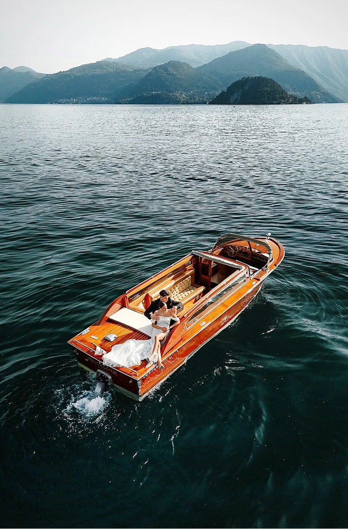 Couple in wedding attire seated on the back of a wooden motorboat on a lake with mountains in the background.