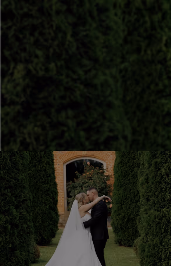 Bride and groom embracing and kissing outdoors between tall green hedges in front of a brick archway.