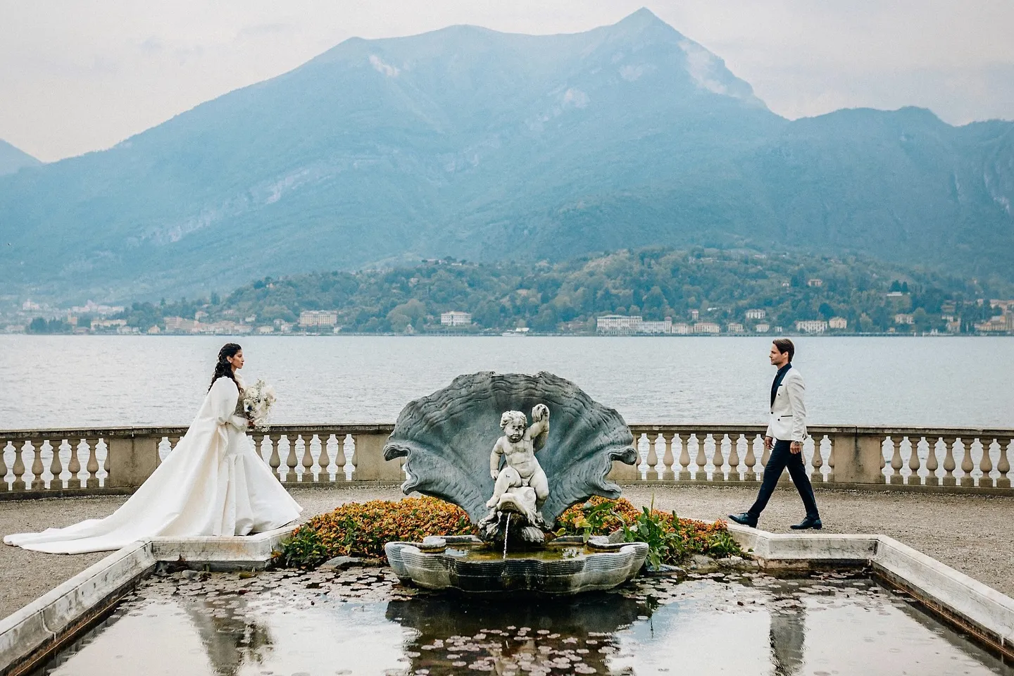 Bride in a white gown and groom in a white jacket walking toward each other near a decorative fountain by a lake with mountains in the background.